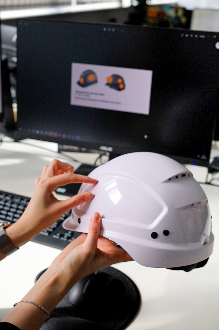 Hände halten einen Arbeitsschutzhelm in die Höhe und zeigen auf eine bestimmte Stelle auf dem Helm.