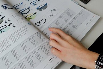 Eine Hand zeigt in einem Brillenkatalog auf eine bestimmte Stelle.