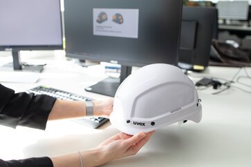 Eine Frau hält an ihrem Schreibtisch einen Arbeitsschutzhelm in den Händen.