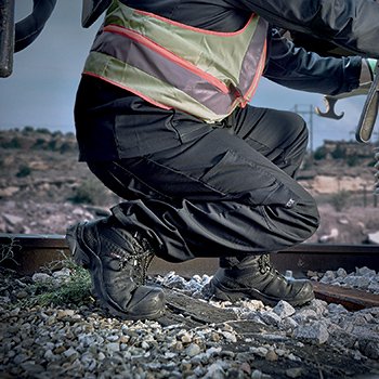 Une personne accroupie vue de côté sur des rails et cailloux, avec des chaussures de sécurité MACCROSSROAD